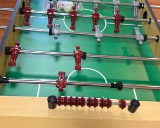 Foosball Table Basement Room — Classic foosball table with a light wood finish and black legs, designed for recreational play typically found in a basement room. It features red and silver player figures mounted on sturdy metal rods with plastic handles, set on a green playfield with white markings. The table includes built-in scoring sliders on both ends for easy score tracking. Measuring approximately 36 inches high, 56 inches wide, and 30 inches deep, it offers a durable and practical game setup. The table is in good condition with minor signs of use but fully functional.