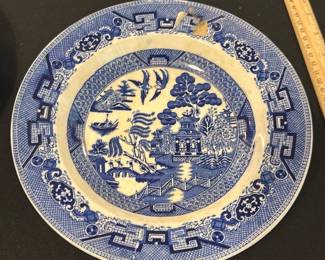Semi China Blue Willow Decorative Plate — This Semi China decorative plate from England features the iconic Blue Willow pattern, showcasing a traditional Asian-inspired design with pagodas, trees, boats, and a bridge in vibrant blue on a cream background. The plate measures approximately 10.5 inches in diameter with a subtle raised rim and is crafted from semi-porcelain china. It exhibits light crazing and one visible chip on the rim, consistent with its age, adding to its vintage character. This collectible piece exemplifies mid-20th-century English ceramic artistry and makes a charming addition to any blue dishes collection or decorative display.