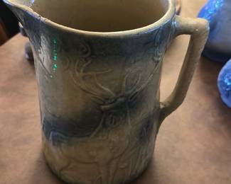 Stoneware Blue Deer Pitcher — This stoneware pitcher features a charming blue and beige glaze with a detailed deer relief design, perfect for rustic or lodge-style dining decor. The pitcher has a sturdy handle and a wide spout for easy pouring. It measures approximately 7 inches tall with a 5-inch diameter. The item is in good condition but does have a small chip on the spout edge. Ideal for collectors of animal motif ceramics or for adding a natural touch to your dining room.