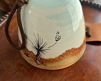 Hand-Painted Ceramic Hanging Bell — This charming hand-painted ceramic hanging bell features a natural scene with dandelion-like floral motifs accented by a light blue and sandy brown glaze. Finished with a sturdy brown cord for hanging, the bell's underside includes a decorative scalloped ceramic clapper. Measuring approximately 5 inches in height and 4 inches in diameter, it makes a delightful decor accent for both indoor and outdoor spaces. The bell is in good condition with minor wear consistent with age and gentle use. Its artisanal style adds a rustic and natural element to any bonus up room or garden setting.