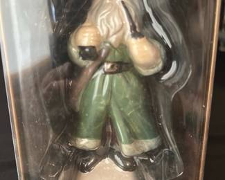Mistletoe Mountain Santa Figurine — This Peebles 1988 Santa figurine from the Traditions of Santa collection is a numbered holiday collectible produced exclusively for Peebles. The ceramic/porcelain figure shows Santa in a green outfit holding a pipe and staff and is presented in its original clear display box with gold backing and snowflake graphics. The boxed figure measures approximately 5.5 inches tall and is a limited-edition run (numbered to 6000 with one-year production notes on the box). In good vintage condition with the figure appearing intact and the box showing light shelf wear and scuffs consistent with age and storage.