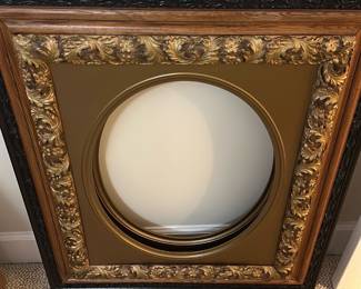 Ornate Gold Leaf Frame with Round Openin — This ornate decorative frame features intricate gold leaf detailing with scrolling acanthus leaves on the inner border, surrounded by a warm wood outer border and accented with black trim. Designed to hold a circular artwork or mirror, it brings a classical and elegant touch to any bedroom or living space. The frame measures approximately 24 inches by 24 inches square with a 12 inch diameter round opening. It is in good condition with minor surface wear consistent with age. Ideal for vintage-style decor or as a statement piece in refined interiors.