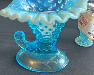 Fenton Blue Opal Hobnail Cornucopia — This charming Fenton blue hobnail cornucopia features iridescent opal glass with a ruffled edge and a distinctive curled tail design. Crafted by Fenton in their signature hobnail pattern, this piece showcases exquisite craftsmanship and vibrant blue coloring with subtle opalescence. Measuring approximately 5.5 inches tall, 6 inches wide, and 4 inches deep, it makes a striking decorative accent for any collection or display. The cornucopia is in good condition with minor surface wear consistent with age and no chips or cracks. A lovely example of vintage Fenton glass, perfect for collectors of hobnail or opal glassware.