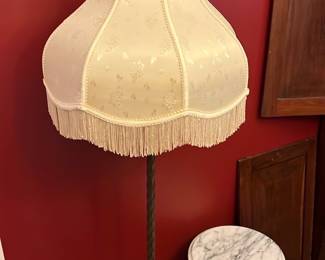 Vintage Brass Floor Lamp w/ Fringe Shade — This vintage brass floor lamp features an ornate twisted pole and elaborately designed base with intricate floral details. The cream fabric lampshade is adorned with subtle floral patterns and a decorative fringe trim, adding a touch of classic elegance. Standing approximately 59 inches tall, this lamp complements traditional or antique-style interiors perfectly. It retains good condition with minor wear consistent with age, enhancing its character and charm. A beautiful addition to any front room décor, this lamp brings both style and warmth.