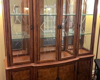Curved Glass China Cabinet — This elegant walnut china cabinet features a curved glass front with etched decorative patterns on the doors, perfect for displaying fine china and collectibles. It includes two glass shelves behind the upper doors and storage cabinets below with wood-paneled doors. Measuring approximately 74 inches tall, 60 inches wide, and 18 inches deep, it offers ample display and storage space in a classic style. The cabinet is in good condition with minor surface wear consistent with age and use. This piece makes a refined addition to any dining room setting.