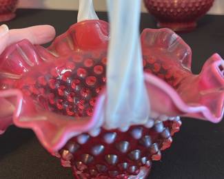 Fenton Cranberry Hobnail Basket — This charming Fenton cranberry hobnail basket features a distinctive twisted white milk glass handle and ruffled edge. The basket is crafted from rich cranberry glass with a textured hobnail pattern, a classic design hallmark of Fenton. It measures approximately 6 inches tall including the handle, with a 5-inch width across the top and 4 inches deep. The piece is in excellent condition with no chips or cracks visible, preserving its vintage charm. A perfect collectible for fans of Fenton glass or elegant vintage home decor.