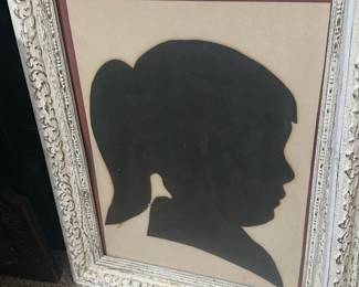 Framed Girl Silhouette Portrait — Framed silhouette portrait of a young girl in a distressed white carved frame, a classic decorative piece for a hallway or bonus room. The silhouette is cut from dark paper and double-matted with a thin burgundy/maroon mat and a beige background as shown. Overall size is estimated and suitable for tabletop or wall display; frame shows intentional distressing and vintage character. In good condition with light surface wear to the frame and glass consistent with age; silhouette paper appears intact with slight fading at edges.