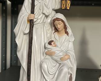 Nativity Holy Family Statue — Porcelain-style Holy Family nativity statue depicting Joseph standing with a staff and lantern, Mary seated cradling the swaddled baby Jesus. Finished in white robes with painted hair, skin tones and a painted brown base; lightweight resin/ceramic construction suitable for tabletop or mantel display. Measures approximately 11 in tall x 6 in wide x 5 in deep, making it an easy, decorative addition to a holiday or year-round devotional display. Good overall condition with minor surface wear to paint on the base and edges consistent with normal display use.