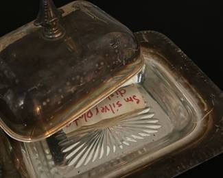 San Silver Plated Butter Dish — This San silver plated butter dish features a classic design with a decorative finial on the lid and a glass base with a cut pattern. The silver plating adds a timeless elegance to this practical tableware item. The dish measures approximately 2.5 inches in height, 6 inches in width, and 5 inches in length. It is in good condition with some tarnish consistent with age but no noticeable dents or damage. A functional and elegant piece perfect for formal dining or vintage table settings.