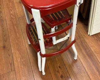Vintage Red and White Metal Step Stool — This vintage metal step stool features a vibrant red seat and steps with a contrasting white frame, perfect for adding retro charm to your basement or kitchen. Constructed from sturdy metal, it combines functionality with a nostalgic design, including a small backrest for added support. Measuring approximately 18 inches high, 14 inches wide, and 14 inches deep, it is compact yet practical for reaching high shelves. The stool shows normal wear and some chipping paint consistent with age but remains solid and stable for use. A charming addition for collectors or practical use in home and garden settings.