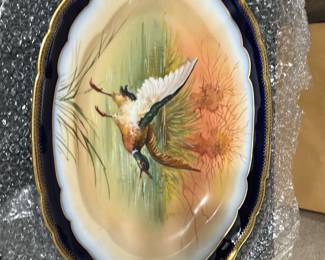 Limoges Hand Painted Duck Platter — This beautiful Limoges hand-painted platter features a detailed duck in flight, surrounded by natural habitat elements. The piece is accented with rich cobalt blue and gilded handles, showcasing fine porcelain craftsmanship from Limoges, France. Measuring approximately 14.5 inches in length and 8.5 inches in width, it offers both decorative appeal and functional serving use. The platter is in good condition with minor surface wear consistent with age, and the hand-painted artistry remains vibrant. Marked with the Limoges France stamp, this platter is a fine example of traditional French porcelain.