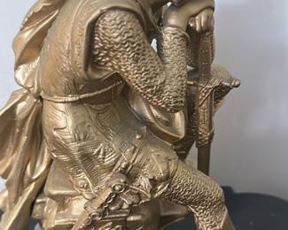 Antique Black & Gold Figural Mantel Cloc — This antique black mantel clock features an ornate gold figural statue of a medieval knight resting on top, showcasing exquisite craftsmanship and detailed design. The clock face is white with black Arabic numerals surrounded by a decorative gold bezel, encased in a black body with intricate gold scrollwork accents. Measuring approximately 15 in height, 16 in width, and 7.5 in depth, it is a substantial decorative piece designed for mantel or shelf display. The clock includes a winding key and appears to be in good condition with minor wear consistent with age but no major damage visible. This elegant piece embodies Victorian or French Empire style, perfect for collectors of vintage timepieces or antique home decor.