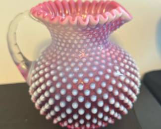 Cranberry Fenton Hobnail Glass Pitcher — This charming cranberry Fenton hobnail glass pitcher showcases a ruffled top and textured hobnail pattern. Crafted from vibrant pink glass with opaque white hobnail bumps, it features a curved handle and a flared spout perfect for decorative or practical use. Measuring approximately 6 inches tall and 6 inches wide, this piece adds vintage elegance to any master bedroom or display. The pitcher is in good condition with no visible chips or cracks, maintaining its glossy finish. A lovely example of classic Fenton artistry, this pitcher makes a delightful collectible.