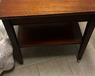 Wood End Table with Lower Shelf — Simple wooden end table with a lower shelf offers practical bedside or living-room use. Constructed in a dark stained wood with a rectangular top and straight square legs, the surface shows light scratches and wear consistent with regular use. Estimated dimensions make it a compact piece suitable for small spaces and it includes small caster feet on two legs for easy repositioning. Overall condition is fair with visible surface wear and finish loss on the top and edges but structurally sound.