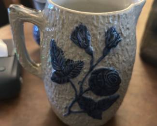 Blue Floral Ceramic Pitcher — This charming ceramic pitcher features a textured off-white base adorned with blue floral designs in relief. The piece includes a sturdy handle and a well-formed spout for easy pouring, marked with the number '18' on the base. Standing approximately 6 inches tall and 5 inches wide, this pitcher is ideal for serving beverages or as a decorative accent in your dining room. The condition is good with slight wear consistent with age but no visible chips or cracks. Its rustic farmhouse appeal makes it a lovely vintage addition to any collection or table setting.