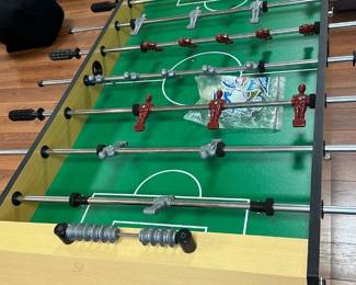 Foosball Table Basement Room — Classic foosball table with a light wood finish and black legs, designed for recreational play typically found in a basement room. It features red and silver player figures mounted on sturdy metal rods with plastic handles, set on a green playfield with white markings. The table includes built-in scoring sliders on both ends for easy score tracking. Measuring approximately 36 inches high, 56 inches wide, and 30 inches deep, it offers a durable and practical game setup. The table is in good condition with minor signs of use but fully functional.
