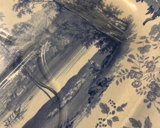 Davenport Blue and White Platter — This Davenport blue and white porcelain platter features a detailed pastoral scene surrounded by an ornate floral border in shades of blue. The scalloped edges add a delicate, vintage charm to this decorative serving piece. Measuring 13.5 inches in width, it provides an elegant accent for any table or display. The platter is in good condition with minor wear and expected age-related surface scratches. Marked on the back with the Davenport brand, it represents English ceramic artistry from the 19th century.