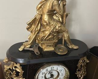Antique Black & Gold Figural Mantel Cloc — This antique black mantel clock features an ornate gold figural statue of a medieval knight resting on top, showcasing exquisite craftsmanship and detailed design. The clock face is white with black Arabic numerals surrounded by a decorative gold bezel, encased in a black body with intricate gold scrollwork accents. Measuring approximately 15 in height, 16 in width, and 7.5 in depth, it is a substantial decorative piece designed for mantel or shelf display. The clock includes a winding key and appears to be in good condition with minor wear consistent with age but no major damage visible. This elegant piece embodies Victorian or French Empire style, perfect for collectors of vintage timepieces or antique home decor.
