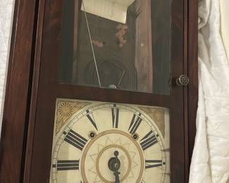Wood Cased Wall Clock — Wood-cased pendulum wall clock with painted metal dial and Roman numerals; photographed in the bedroom up room. The case is a dark wood finish with a glass door showing the clock face and upper compartment (pendulum and weights removed or stored inside). Overall measurements estimated for a wall regulator-style clock; face shows wear and some paint loss, case has surface scratches and a missing lower molding piece. Measuring approximately 36 in tall and 14 in wide, depth about 6 in, the clock is sold as a decorative vintage/estate piece and may need clockmaker service to run. Good cosmetic condition for display with expected wear for age and small losses to finish and dial.
