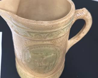 Vintage Ceramic Pitcher with Cow Motif — This vintage ceramic pitcher features a glaze with a distinctive raised cow motif on its side. The durable ceramic construction includes a textured handle and a patterned band encircling the top and bottom edges. Measuring approximately 6.5 inches tall with a 4.5 inch width, it is ideal for decorative use or serving liquids. The pitcher is in good condition with some minor surface wear consistent with age, and no visible cracks or chips. Its charming pastoral design makes it a quaint addition to farmhouse or country-style decor.