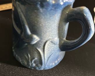 Blue Stoneware Mini Mug — This charming blue stoneware mini mug features a textured floral and leaf relief design that adds a rustic and handcrafted appeal. The mug is compact and solidly built, perfect as a decorative piece or for serving small beverages or a test cup. Its rich blue glaze complements the intricate detailing and gives it an artisanal feel. The mug is in good condition with minor surface imperfections consistent with handcrafted pottery. Ideal for collectors of handmade ceramics or as a unique accent piece in gift shelves or kitchen displays.