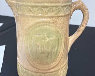 Vintage Ceramic Pitcher with Cow Motif — This vintage ceramic pitcher features a glaze with a distinctive raised cow motif on its side. The durable ceramic construction includes a textured handle and a patterned band encircling the top and bottom edges. Measuring approximately 6.5 inches tall with a 4.5 inch width, it is ideal for decorative use or serving liquids. The pitcher is in good condition with some minor surface wear consistent with age, and no visible cracks or chips. Its charming pastoral design makes it a quaint addition to farmhouse or country-style decor.