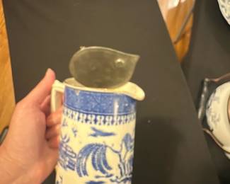 T. G. M. Co. Blue Willow Pitcher — This vintage T. G. M. Co. porcelain pitcher features the classic Blue Willow pattern in blue and white with traditional Asian-inspired scenic designs. It includes a metal hinged lid with a finial and a sturdy handle, perfect for serving beverages. The pitcher measures approximately 7.5 inches in height and 4.5 inches in width, making it a practical and decorative addition to any kitchen or dining collection. The piece is in good condition, displaying minor signs of wear consistent with age, and the metal lid shows some tarnishing. Marked on the base as "T.G.M.Co. Blue Willow," this collectible pitcher embodies the charm of English transferware.