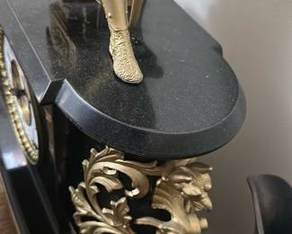 Antique Black & Gold Figural Mantel Cloc — This antique black mantel clock features an ornate gold figural statue of a medieval knight resting on top, showcasing exquisite craftsmanship and detailed design. The clock face is white with black Arabic numerals surrounded by a decorative gold bezel, encased in a black body with intricate gold scrollwork accents. Measuring approximately 15 in height, 16 in width, and 7.5 in depth, it is a substantial decorative piece designed for mantel or shelf display. The clock includes a winding key and appears to be in good condition with minor wear consistent with age but no major damage visible. This elegant piece embodies Victorian or French Empire style, perfect for collectors of vintage timepieces or antique home decor.