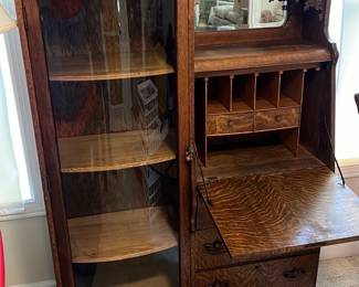 Antique Oak Secretary Curio Cabinet — 41”x68”. This antique oak secretary curio cabinet features intricate carved details and a glass display case. The piece combines functional storage with elegant design, including a fold-down writing surface, small drawers, and cubbies, topped with a decorative mirror and ornate carvings on the crest. Measuring approximately 59 inches tall, 43 inches wide, and 18 inches deep, it offers versatile use for office or living space. The cabinet is in good condition with typical minor wear consistent with age, showcasing the natural oak grain and craftsmanship. This classic piece represents early 20th-century furniture making with quality solid oak construction and distinctive embellishments.