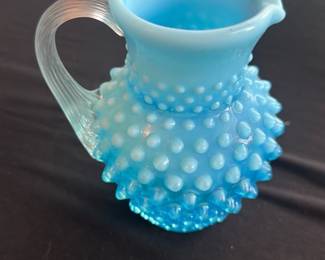 Blue Opalescent Hobnail Glass Pitcher — This charming blue opalescent hobnail glass pitcher features a textured hobnail pattern around the body and a clear ribbed handle. The pitcher has a delicate spout and a graceful silhouette, making it a delightful decorative and functional piece. Measuring approximately 6 inches tall with a width of 4.5 inches, it is a perfect size for serving beverages or as a decorative accent. The glass is in excellent condition with no chips or cracks visible. This piece adds vintage charm and character to any collection or table setting.