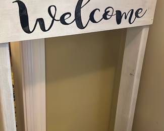 White Wooden Welcome Sign — This white wooden welcome sign features the word 'welcome' in elegant black script across a sturdy frame. Crafted from painted wood, it offers a rustic charm ideal for front room decor or entryways. The piece measures approximately 36 inches wide, 30 inches tall, and 6 inches deep. It is in good condition with minor surface wear visible. This decorative sign adds a warm, inviting touch to any home setting.