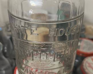 White House Vinegar Glass Bottle — This vintage White House Vinegar glass bottle features embossed lettering and a detailed depiction of the White House. Made of clear glass with a loop handle and cork stopper, it measures approximately 10 inches in height. The bottle is a charming collectible piece ideal for display or decorative use. It is in good condition, with minor wear consistent with age and no visible cracks or chips. This collectible reflects historical Americana branding and makes an excellent addition to vintage glassware collections.