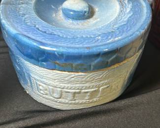 Vintage Blue Ceramic Butter Crock — This vintage blue ceramic butter crock features a textured design with the word 'BUTTER' embossed on the front. It has a metal handle with a distressed wooden grip, adding rustic charm to its classic look. Measuring approximately 4.5 inches tall and 5.5 inches wide with a 4.5-inch diameter, it is ideal for butter storage and countertop display. The crock shows some age-related wear and minor chipping around the rim but remains functional and attractive. Its sturdy construction and charming design make it a collectible piece for any kitchen or farmhouse decor.