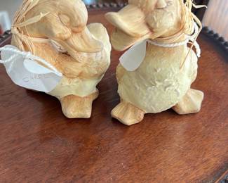 Duck Figurines Set — This charming set of carved wooden duck figurines features a pair of light-colored ducks with detailed feather textures and natural straw accents around their necks. Each figurine displays a rustic, handcrafted appeal, perfect for adding a touch of country or farmhouse decor to any room. Measuring approximately 4 inches tall and 3 inches wide, these lightweight pieces fit easily on shelves or tabletops. Both ducks are in good condition, showing minor wear consistent with handmade wooden decor. Ideal for collectors or as a quaint decorative accent.