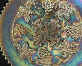 Carnival Glass Compote Plate — This striking Carnival glass compote plate features an iridescent finish with a grape and leaf pattern. The plate showcases a detailed grapevine design with a checked border and crimped edge, typical of mid-20th century Carnival glass styles. Estimated to measure approximately 8 inches in diameter, it serves as a decorative addition to any vintage glass collection or tabletop display. The piece is in good condition with minor surface wear consistent with age and use. Carnival glass is prized for its vibrant colors and collectible appeal.