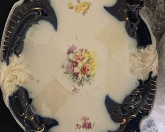 RS Prussia Blue Trim Porcelain Plate — This RS Prussia porcelain plate features a striking blue trim with ornate relief designs and delicate floral patterns in the center. The piece is an elegant example of early 20th century European porcelain craftsmanship. Measuring approximately 10 inches in length and 7.5 inches in width, it is suitable for display or collection. The plate is in good condition with no visible cracks but shows minor age-related wear. Originally made in Prussia, this collectible plate adds vintage charm to any collection.