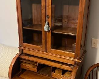 Wood Roll-Top Secretary Desk — Attractive wooden roll-top secretary desk with glass-front upper cabinet and brass hardware; perfect for a master bedroom or home office. Constructed of medium-tone hardwood with a burled roll-top, three lower drawers with decorative brass pulls, and an upper display cabinet with two glass doors and interior shelving. Overall footprint is medium-sized and practical for writing and storage; roll-top opens to reveal small cubbies and drawers for organization. Good estate-sale condition with light surface wear and patina consistent with normal use, hardware intact and drawer function smooth.