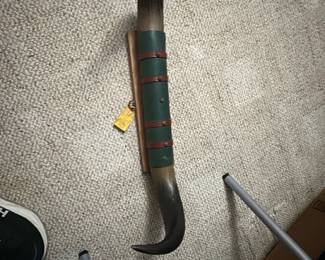 Danny Ballard 1960s Horn Display — This large decorative horn, gifted from Danny Ballard circa early 1960s at the Memphis Fair, offers a unique vintage touch for any collection or rustic decor. The piece features authentic horn with green and brown leather strap detailing, mounted on a wooden stand for display. Measuring approximately 48 inches in length with a curved and pointed shape, this horn is a striking accent piece for a bonus room or man cave. It is in good condition with natural wear consistent with its age. This collectible represents mid-20th-century Americana and is a notable conversation starter.