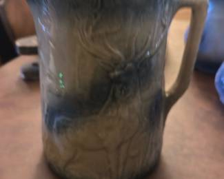 Stoneware Blue Deer Pitcher — This stoneware pitcher features a charming blue and beige glaze with a detailed deer relief design, perfect for rustic or lodge-style dining decor. The pitcher has a sturdy handle and a wide spout for easy pouring. It measures approximately 7 inches tall with a 5-inch diameter. The item is in good condition but does have a small chip on the spout edge. Ideal for collectors of animal motif ceramics or for adding a natural touch to your dining room.