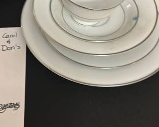 Noritake-Derry White China Set — This elegant Noritake-Derry china set features classic white porcelain with delicate silver rims, perfect for formal dining. The set includes a large oval platter, nine large dinner plates, two soup bowls, eight salad plates, eight cups, eight saucers, and additional serving pieces including cream, sugar, salt and pepper set, butter dish with lid, two oval vegetable bowls, and one divided vegetable bowl. The pristine white finish with silver accents adds a timeless sophistication to any table setting. All pieces are in good condition with minor signs of use consistent with careful everyday dining. Ideal for collectors or those who appreciate fine dining ware from the bonus up room collection.