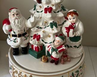 Lenox Holiday Santa's Tree Centerpiece — This charming Lenox Holiday Decorating Santa's Tree Centerpiece features a festive group of Santa Claus and children around a decorated Christmas tree with red bows and gold accents. Handcrafted in China, this ceramic piece boasts exquisite detail and delicate painting, perfect for holiday décor in any home office or living space. Measuring approximately 8 inches high and 8 inches wide, it serves as a beautiful tabletop decoration to bring holiday cheer. The centerpiece is in excellent condition with no visible chips or damage, retaining its original paint and finish. A timeless collectible, this piece is a wonderful addition to any Lenox Christmas collection dating from the early 2000s.