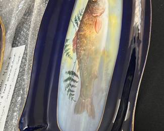 Limpges France Cobalt Blue Fish Platter — This Limpges France fish platter features a striking hand-painted fish design against a cobalt blue background. Crafted from fine porcelain, it showcases intricate naturalistic details and delicate gold trim accents on its ornate edges. Measuring approximately 18 inches in length and 7 inches in width, this platter is perfect for serving or display. It is in excellent condition with no visible chips or cracks, reflecting high-quality craftsmanship. A beautiful example of French porcelain artistry, ideal for collectors or as a decorative piece.