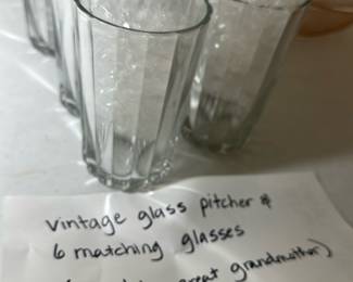 Vintage Glass Pitcher & 6 Glasses — This vintage glass pitcher comes with six matching glasses, each featuring a classic vertical ribbed design. The set is clear and crafted from sturdy glass, making it perfect for serving beverages in style. The pitcher measures approximately 7.5 inches tall and the glasses stand about 5 inches tall. The set is in good condition with no visible chips or cracks and carries sentimental value as it was owned by the seller's great-grandmother.