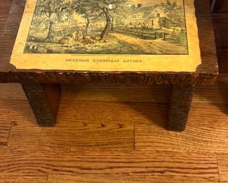 American Homestead Autumn Wooden Stool — This rustic wooden stool features a charming vintage print titled 'American Nostalgia Autumn' on its top surface, giving it a nostalgic farmhouse appeal. The stool is crafted from dark-stained wood with a heavily textured, hand-carved design on the legs and edges, enhancing its handcrafted aesthetic. Measuring approximately 15 inches in height, 20 inches in width, and 11 inches in depth, it is a sturdy accent piece suitable for the living room. The stool is in good condition with minor wear consistent with age and use, maintaining its strong structural integrity. Its quaint design and vintage print make it a unique decorative as well as functional item for country-style or rustic interiors.