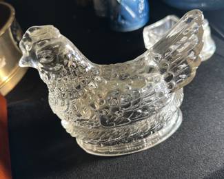 Clear Glass Hen — This clear glass hen covered dish features detailed textured patterns resembling feathers and a woven basket base. The hen serves as a removable lid, revealing a practical storage bowl beneath. Measuring approximately 4.5 inches tall, 7 inches wide, and 5 inches deep, this vintage collectible is perfect for decorative display or practical use. The dish is in good condition with no visible chips or cracks. A classic piece often used in kitchens or dining areas, adding vintage charm to your home.