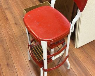 Vintage Red and White Metal Step Stool — This vintage metal step stool features a vibrant red seat and steps with a contrasting white frame, perfect for adding retro charm to your basement or kitchen. Constructed from sturdy metal, it combines functionality with a nostalgic design, including a small backrest for added support. Measuring approximately 18 inches high, 14 inches wide, and 14 inches deep, it is compact yet practical for reaching high shelves. The stool shows normal wear and some chipping paint consistent with age but remains solid and stable for use. A charming addition for collectors or practical use in home and garden settings.