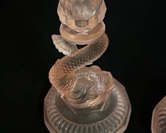 Pink Dolphin Glass Candlestick Holder — This delicate pink glass candlestick holder features a unique dolphin design coiled around the stem, elevating its decorative appeal. The holder combines a flared top for candle placement with a sturdy, detailed base that showcases fine craftsmanship in translucent pink glass. Measuring approximately 7 inches in height and 3 inches in diameter at the base, it fits well as a charming accent piece in any room. The condition is excellent with no visible chips or cracks, maintaining its elegant finish. Its distinctive aquatic motif makes it a standout conversation piece for collectors or decor enthusiasts.