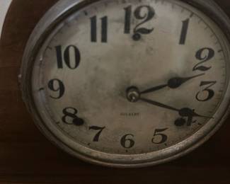 Gilbert Vintage Wooden Mantel Clock — This charming Gilbert vintage wooden mantel clock features a classic tambour style with smooth curved edges. The clock face shows aged patina with bold black numerals and the Gilbert brand mark visible. Measuring approximately 9 inches tall, 17 inches wide, and 5 inches deep, this clock is designed for display on a mantel or shelf. The wood casing exhibits some wear consistent with age but remains structurally sound. A fine example of early 20th-century American clockmaking, it adds vintage character to any room.