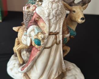 Ceramic Santa with Reindeer musical — This decorative ceramic Santa with reindeer is a charming holiday mantel or tabletop accent captured in soft cream, red and teal tones. The figure shows Santa carrying gifts with textured robe details and a realistic reindeer companion; base has a snowy ground and the underside bears handwritten provenance notes. Overall size is compact for display on shelves or holiday vignettes. Good estate condition with light surface wear; mechanism on base (battery or music box) appears present but untested.