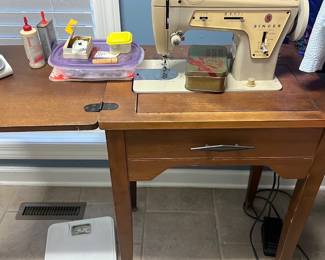 Singer Sewing Machine Cabinet — This classic Singer beige sewing machine is mounted in a wooden sewing table and ready for basic mending and sewing projects. The metal-bodied machine sits in a fold-out tabletop with a single drawer beneath and shows typical surface wear to the cabinet and light cosmetic wear to the machine finish. Overall unit measures approximately 36 in tall (table top to floor), 40 in wide (with leaf extended) and 18 in deep; table is relatively lightweight and the machine is a standard domestic size. Machine appears functional but has not been recently tested; cabinet shows scratches and a missing finish spot on one leg consistent with normal use.